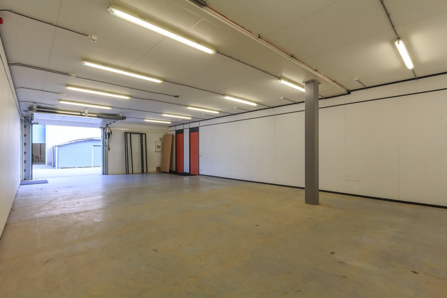 Lege bedrijfsruimte met betonnen vloer, witte wanden, TL-verlichting en een open garagedeur.